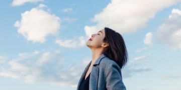 600700882ad8e Portrait of positive young Asian woman with eye closed, enjoying sunlight under blue sky and clouds.