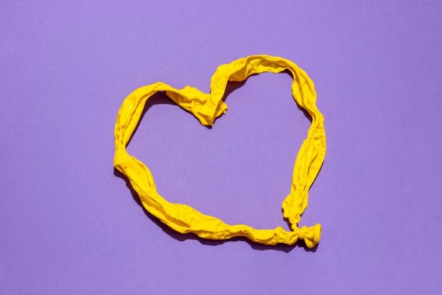 Elevated view of heart shaped deflated yellow balloon against purple background. Relationship breakup concept.