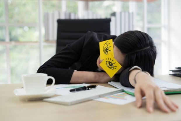 Young business woman with fake eyes painted on adhesive note sleeping at workplace in office
