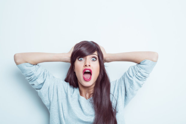 Portrait of shocked young woman wearing grey jumper, raising her arms and shouting at camera, rolling eyes. Studio shot, white background.