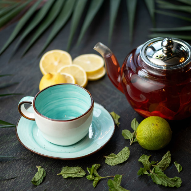 Full teapot with lemon and mint Black tea and empty cup on dark table background. Lime, lemon slices and fresh green mint leaves on it. Soft focus.
