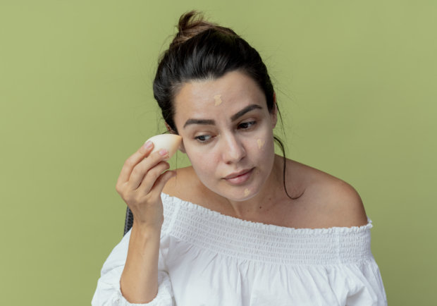 pleased beautiful girl applying foundation with sponge isolated on green background with copy space